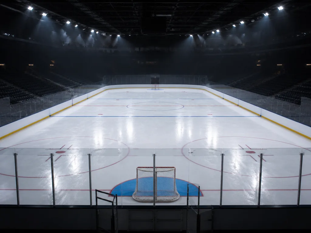 Ishockeyrink med blå och röda linjer på vit is, målbur i förgrunden och dramatisk arenabelysning ovanifrån